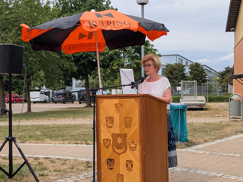 Frau mit Brille und weißem Oberteil steht am Rednerpult aus Holz mit 9 verschiedenen Wappen an der Front, mit Mikrofon unter schwarz-orangem Sonnenschirm mit der Aufschrift "Kolping", im Freien.