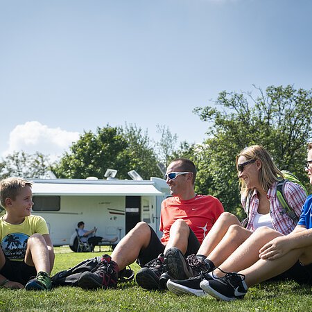 Vier Personen sitzen auf einer Wiese vor Wohnmobilstellplatz in Monheim, im Hintergrund ist ein Wohnmobil, Bäume und blauer Himmel.