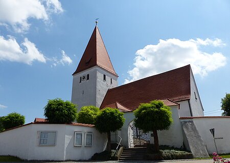 Kirche mit rotem Dach, Turmuhr und umgebender weißer Mauer mit Eingangstor bei blauem Himmel und Bäumen davor.