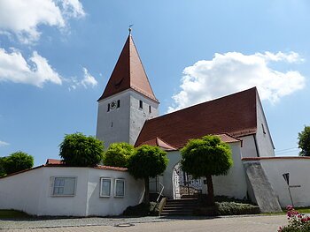 Kirche mit rotem Dach, Turmuhr und umgebender weißer Mauer mit Eingangstor bei blauem Himmel und Bäumen davor.