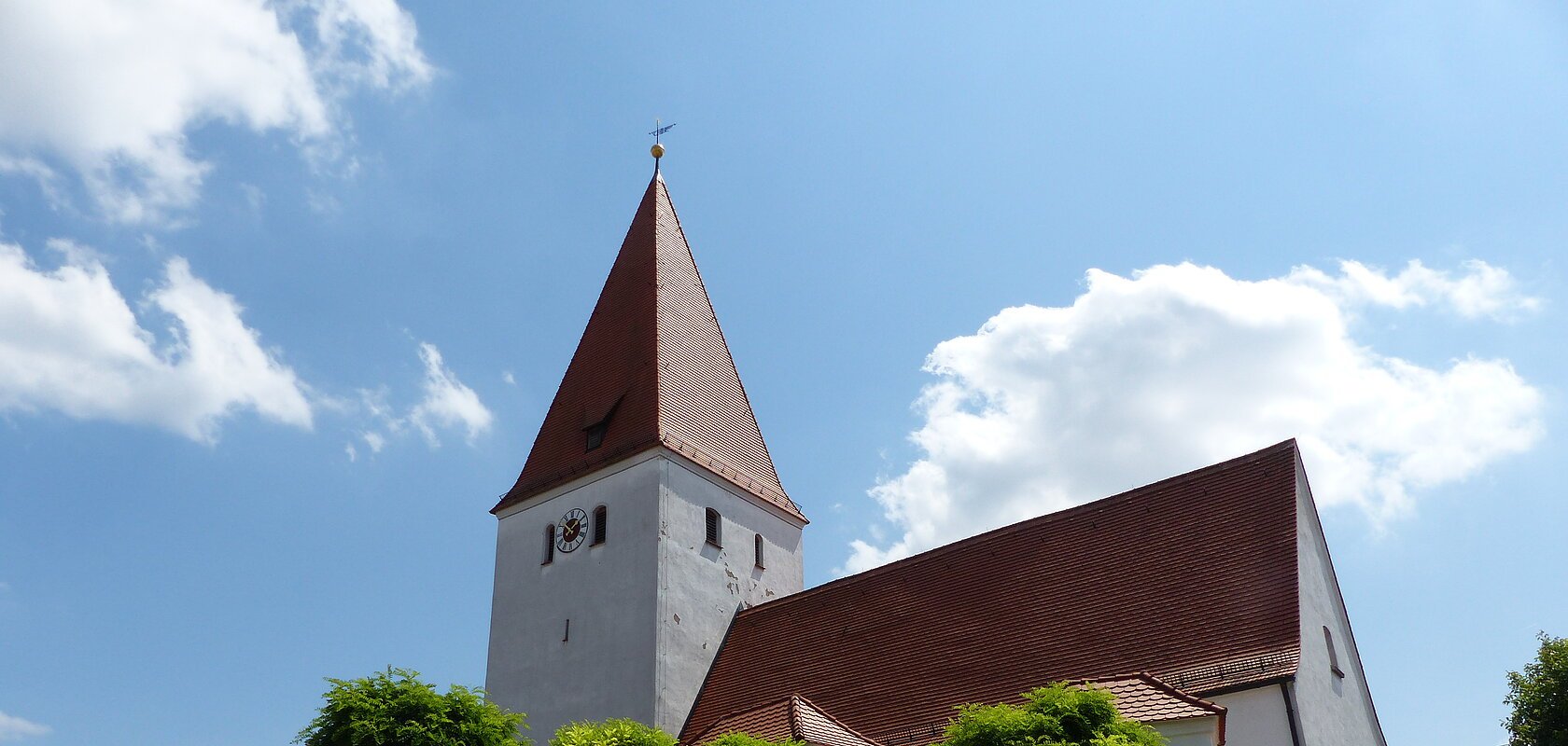 Kirche mit rotem Dach, Turmuhr und umgebender weißer Mauer mit Eingangstor bei blauem Himmel und Bäumen davor.