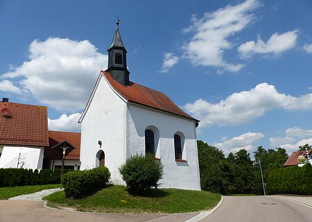 Kleine weiße Filialkirche mit rotem Dach und kleinem Turm oben drauf an einer Straße bei blauem Himmel mit Wolken. Umgeben von grüner Wiese und Hecken, daneben ein Jesuskreuz aus Holz. Im Hintergrund weiteres weißes Haus zu sehen.