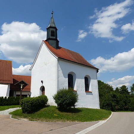 Kleine weiße Filialkirche mit rotem Dach und kleinem Turm oben drauf an einer Straße bei blauem Himmel mit Wolken. Umgeben von grüner Wiese und Hecken, daneben ein Jesuskreuz aus Holz. Im Hintergrund weiteres weißes Haus zu sehen.