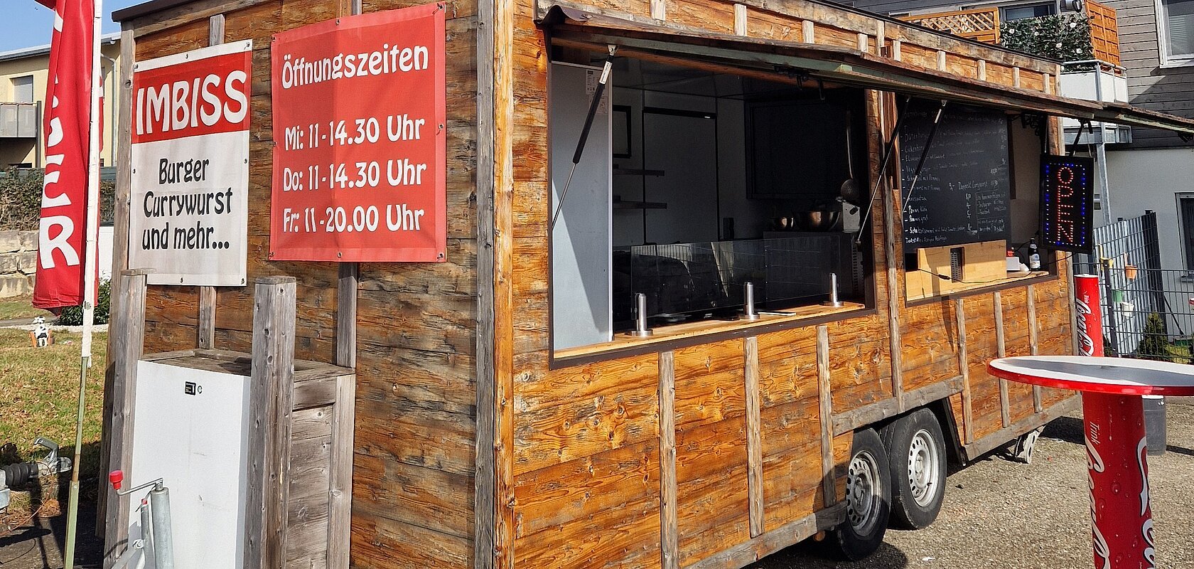 Holz-Imbisswagen mit geöffnetem Fenster, roten Schildern mit Speisen und Öffnungszeiten, stehend auf Asphalt bei blauem Himmel und einem weiteren Haus im Hintergrund.