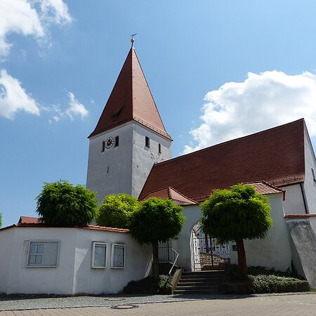 Kirche mit rotem Dach, Turmuhr und umgebender weißer Mauer mit Eingangstor bei blauem Himmel und Bäumen davor.