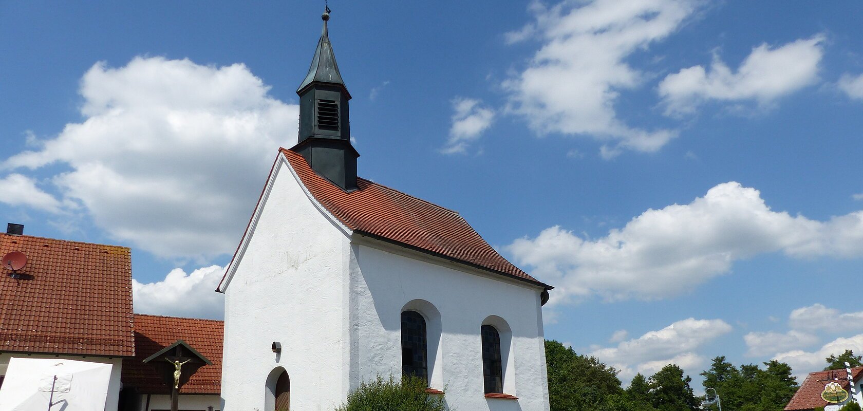 Kleine weiße Filialkirche mit rotem Dach und kleinem Turm oben drauf an einer Straße bei blauem Himmel mit Wolken. Umgeben von grüner Wiese und Hecken, daneben ein Jesuskreuz aus Holz. Im Hintergrund weiteres weißes Haus zu sehen.