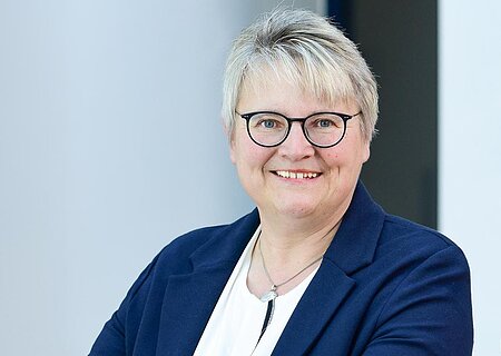 Porträt einer lächelnden Frau mit kurzen grauen Haaren, Brille, weißem Hemd und blauem Blazer, die an einem Geländer lehnt.
1. Bürgermeisterin der Stadt Monheim