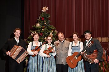 Sechs Personen in Trachten vor geschmücktem Weihnachtsbaum mit Musikinstrumenten posieren vor rotem Vorhang. Heilige Nacht mit Pfarrer R.M. Schießler & Familienmusik Rehm & Rupert Biegel am 21.12.2026 in der Monheimer Stadthalle