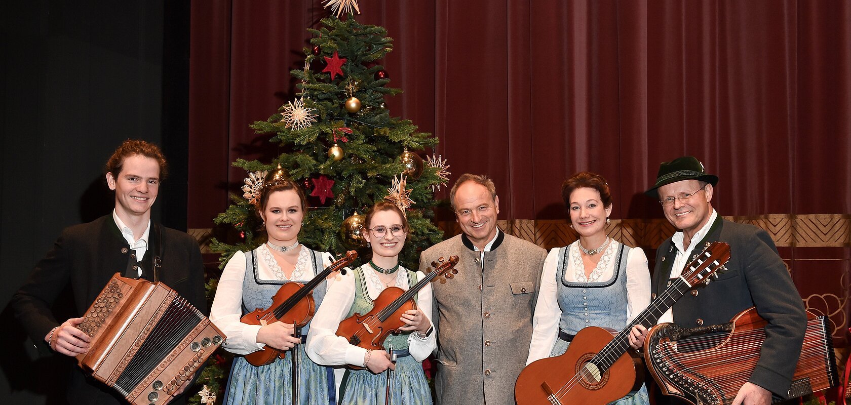 Sechs Personen in Trachten vor geschmücktem Weihnachtsbaum mit Musikinstrumenten posieren vor rotem Vorhang. Heilige Nacht mit Pfarrer R.M. Schießler & Familienmusik Rehm & Rupert Biegel am 21.12.2026 in der Monheimer Stadthalle