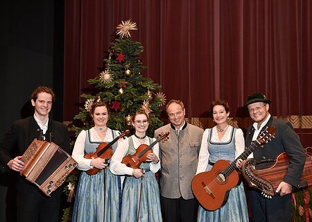 Sechs Personen in Trachten vor geschmücktem Weihnachtsbaum mit Musikinstrumenten posieren vor rotem Vorhang. Heilige Nacht mit Pfarrer R.M. Schießler & Familienmusik Rehm & Rupert Biegel am 21.12.2026 in der Monheimer Stadthalle
