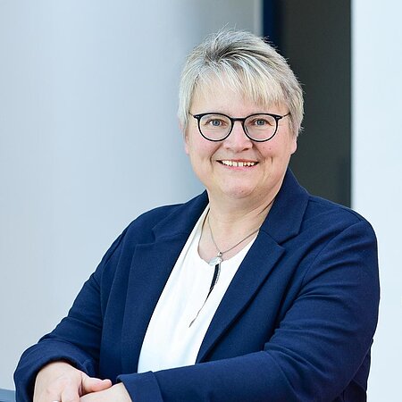 Porträt einer lächelnden Frau mit kurzen grauen Haaren, Brille, weißem Hemd und blauem Blazer, die an einem Geländer lehnt.
1. Bürgermeisterin der Stadt Monheim