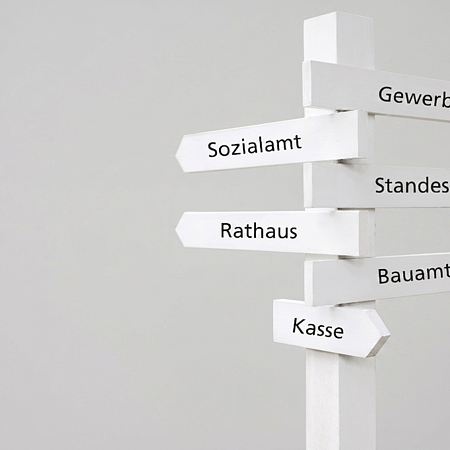 Wegweiser mit weißen Schildern zu Gewerbeamt, Sozialamt, Standesamt, Rathaus, Bauamt und Kasse vor grauem Hintergrund.
