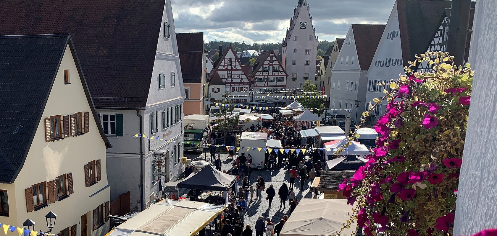 Ansicht von oben auf die Monheimer Innenstadt mit Marktständen links und rechts und Menschen, umgeben von Fachwerkhäusern. Im Vordergrund und an der Hausfassade pinke Blumen.