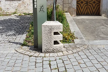 Steinskulptur des Buchstabens "E", dahinter ein Informationsschild und ein Baum mit einem dahinter liegendem Beet. Umgeben von einem gepflastertem Platz, mit einem historischen Gebäude im Hintergrund.
