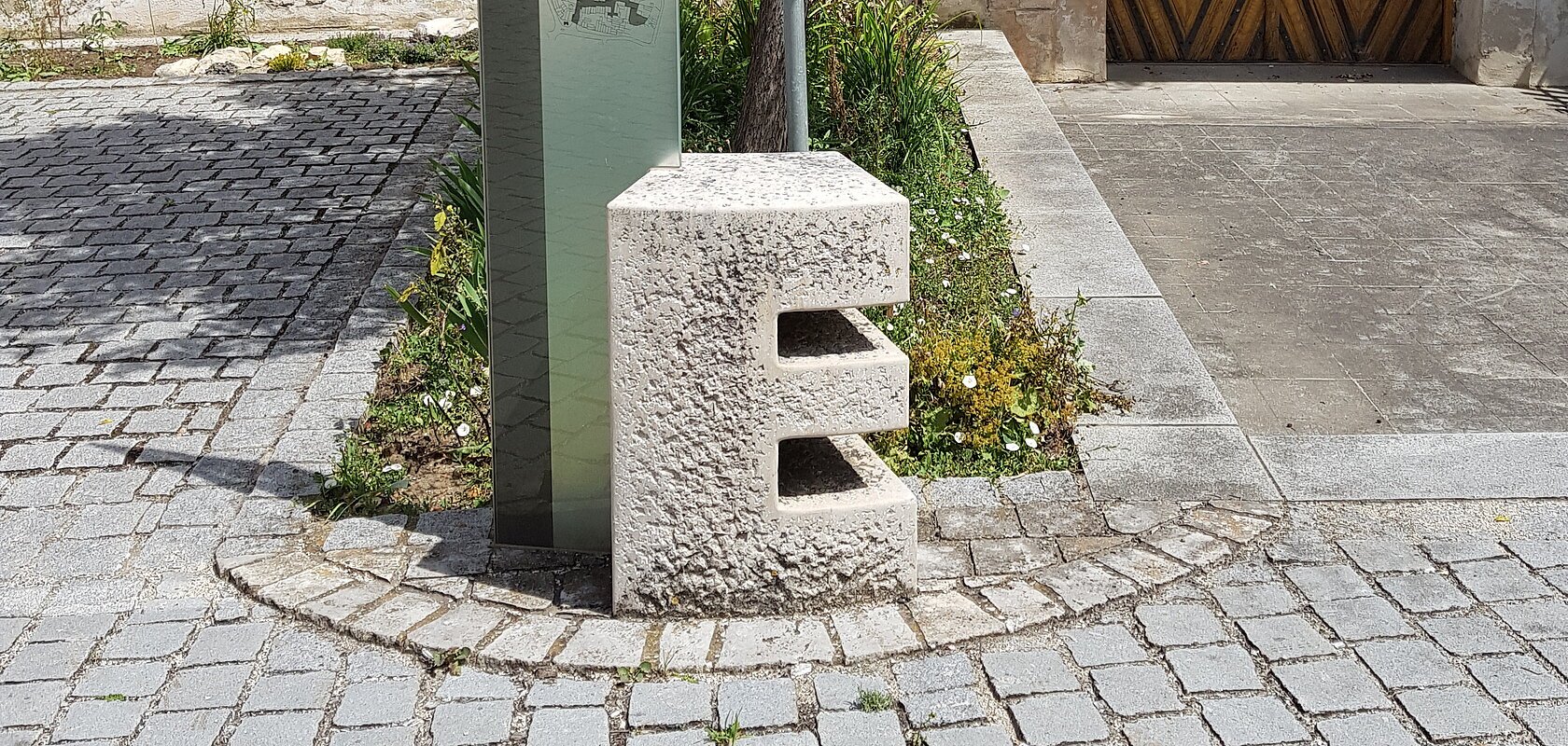 Steinskulptur des Buchstabens "E", dahinter ein Informationsschild und ein Baum mit einem dahinter liegendem Beet. Umgeben von einem gepflastertem Platz, mit einem historischen Gebäude im Hintergrund.