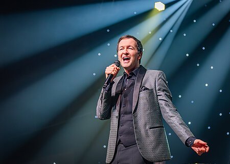 Udo Jürgens - Das Konzert mit Alex Parker & Special Guest Mann im Anzug singt mit Mikrofon auf Bühne vor beleuchtetem Hintergrund mit Sternenlicht.
Udo Jürgens - Das Konzert mit Alex Parker & Special Guest am 20.02.2027 in der Monheimer Stadthalle.