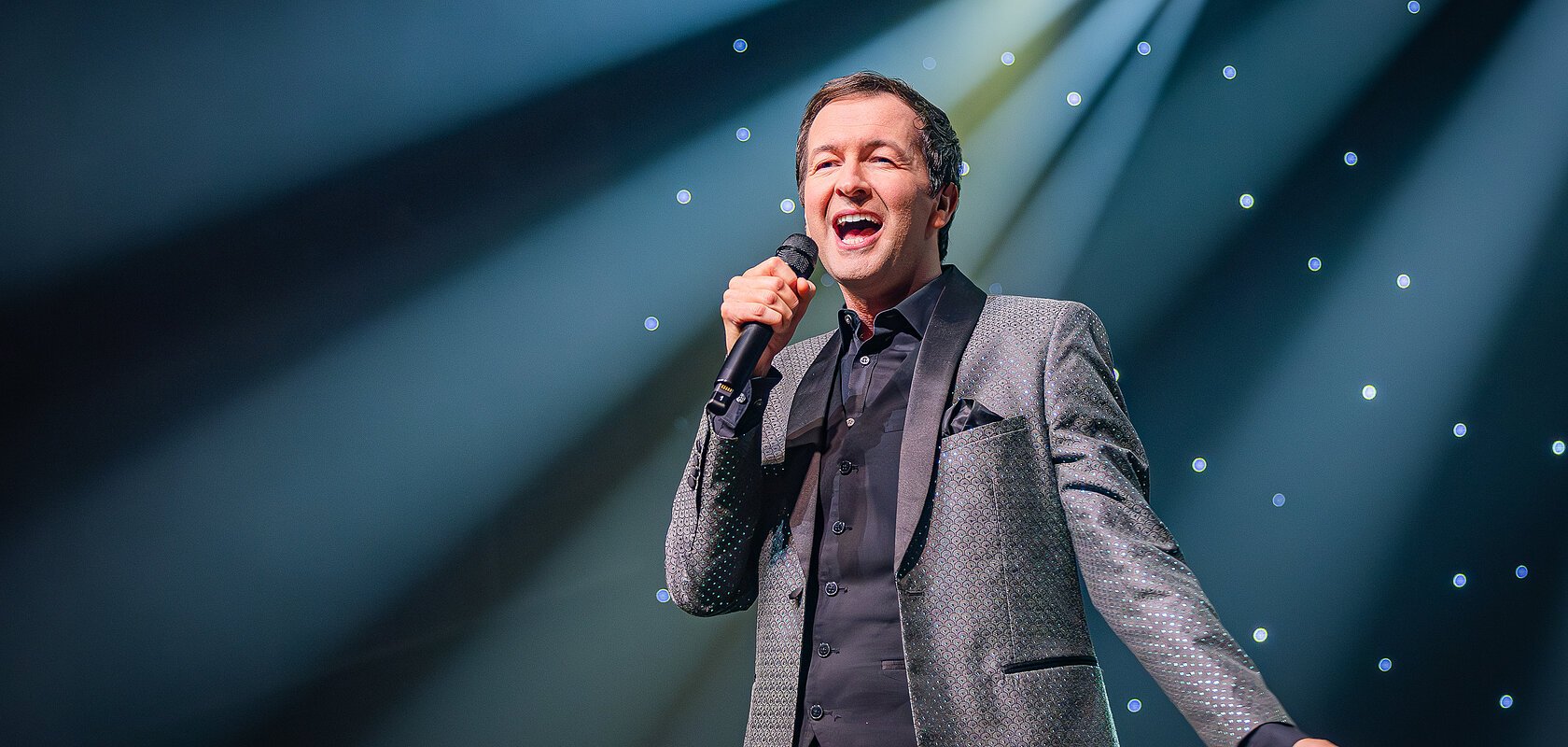 Mann im Anzug singt mit Mikrofon auf Bühne vor beleuchtetem Hintergrund mit Sternenlicht.
Udo Jürgens - Das Konzert mit Alex Parker & Special Guest am 20.02.2027 in der Monheimer Stadthalle.