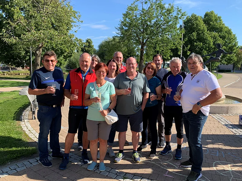 Gruppe von elf Erwachsenen umgeben von grüner Wiese rechts steht auf einem gepflastertem Platz, hält Getränke in der Hand. Bäume, blauer Himmel und Brunnen im Hintergrund.