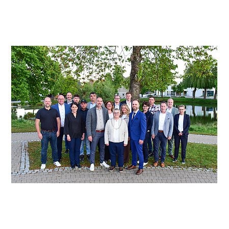 Gruppenfoto von 18 Personen vor einem Teich mit Bäumen und Gebäuden im Hintergrund. Stadtrat Mitglieder der Stadt Monheim - Stand Mai 2026