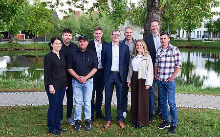 Neun Personen stehen vor einem Baum und einem Teich auf einer Wiese, im Hintergrund Gebäude und Bäume. Die neu gewählten Stadtrat-Mitglieder der Stadt Monheim - im Mai 2026