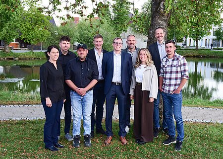 Neu gewählte Stadtrat-Mitglieder Stadt Monheim - Mai 2026 Neun Personen stehen vor einem Baum und einem Teich auf einer Wiese, im Hintergrund Gebäude und Bäume. Die neu gewählten Stadtrat-Mitglieder der Stadt Monheim - im Mai 2026