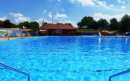 Das Freibad in Monheim: Schwimmbecken mit klarem Wasser, umgeben von Liegestühlen, einem roten Gebäude und Umkleidekabinen im Hintergrund.