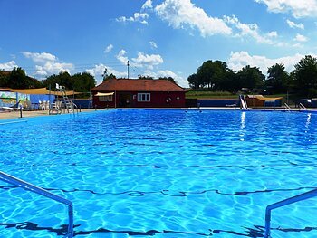 Das Freibad in Monheim: Schwimmbecken mit klarem Wasser, umgeben von Liegestühlen, einem roten Gebäude und Umkleidekabinen im Hintergrund.
