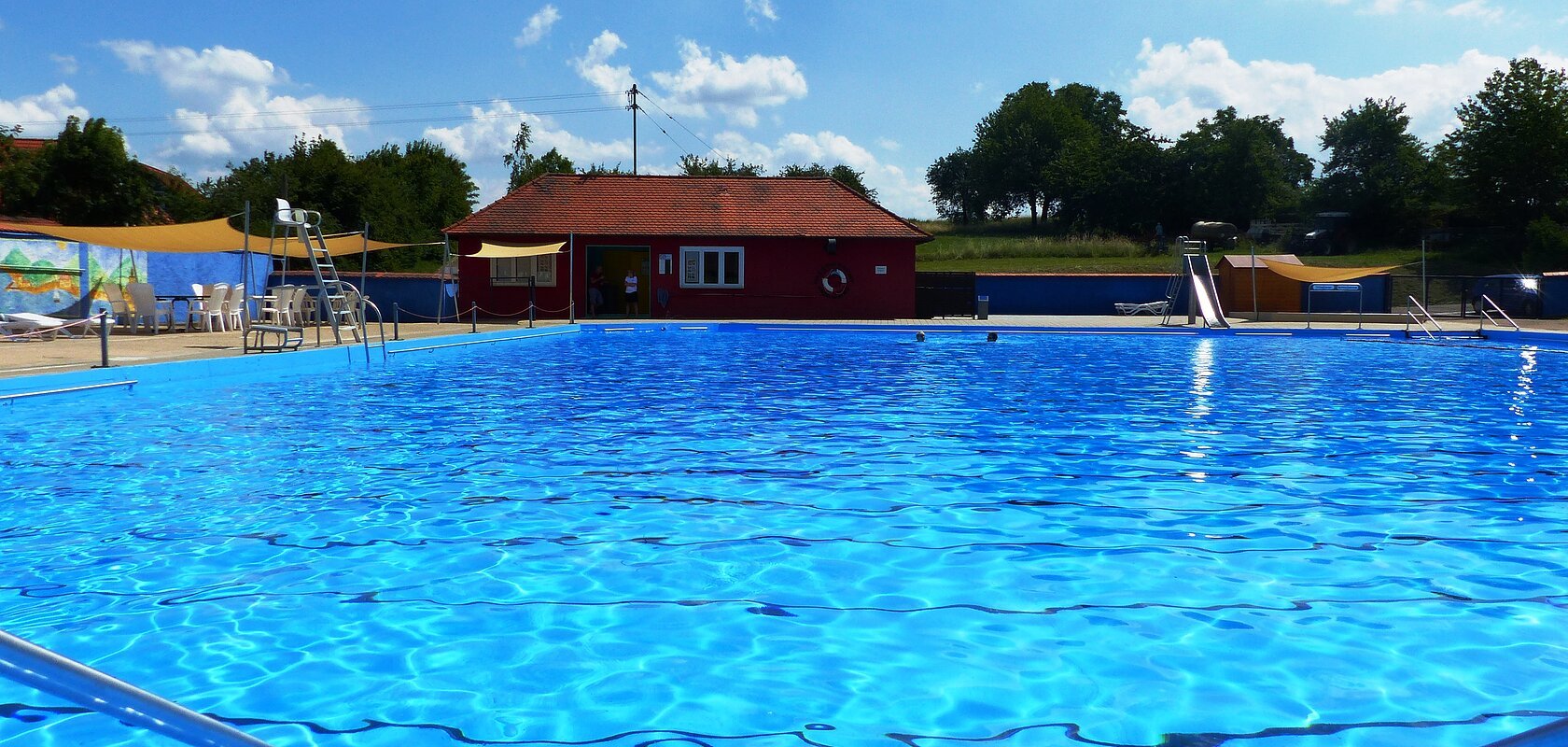 Das Freibad in Monheim: Schwimmbecken mit klarem Wasser, umgeben von Liegestühlen, einem roten Gebäude und Umkleidekabinen im Hintergrund.