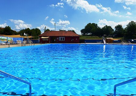 Freibad Monheim - Nostalgiebad mit umfassendem Angebot Das Freibad in Monheim: Schwimmbecken mit klarem Wasser, umgeben von Liegestühlen, einem roten Gebäude und Umkleidekabinen im Hintergrund.
