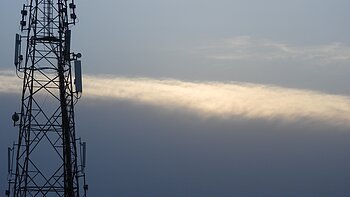 Mobilfunk-Sendemast vor bewölktem Himmel mit einer schmalen, hellen Wolke im Hintergrund.