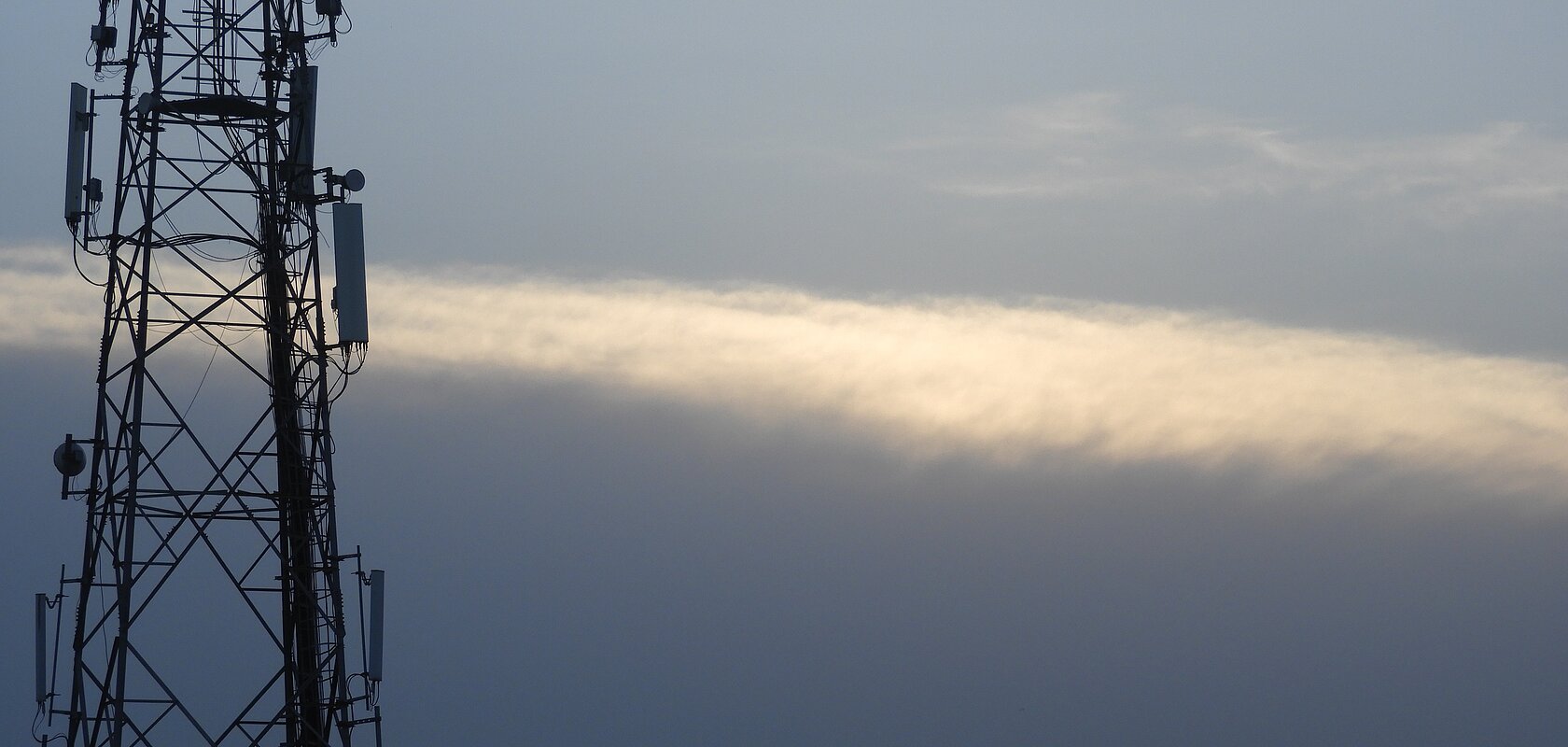Mobilfunk-Sendemast vor bewölktem Himmel mit einer schmalen, hellen Wolke im Hintergrund.