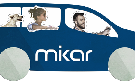 Blaues Auto mit zwei Personen und einem Hund, auf dem Auto der Schriftzug "mikar" auf der Seite. Fahrer hält das Lenkrad.