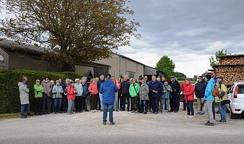 Gruppe von etwa 30 Personen steht auf einem Hof vor Scheunen und Holzstapel, eine Person spricht zur Gruppe. Senioren wandern mit SoMit am 13. Mai 2026 unterwegs in Sulzdorf