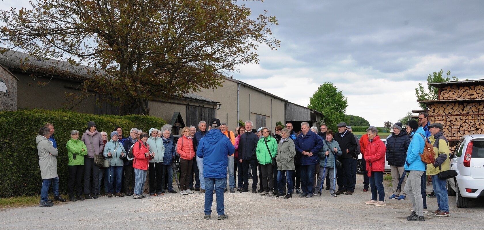 Gruppe von etwa 30 Personen steht auf einem Hof vor Scheunen und Holzstapel, eine Person spricht zur Gruppe. Senioren wandern mit SoMit am 13. Mai 2026 unterwegs in Sulzdorf