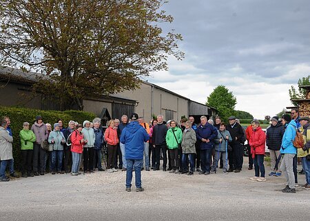 SoMit Wanderung 13.05.2026 in Sulzdorf Gruppe von etwa 30 Personen steht auf einem Hof vor Scheunen und Holzstapel, eine Person spricht zur Gruppe. Senioren wandern mit SoMit am 13. Mai 2026 unterwegs in Sulzdorf