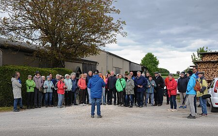 Gruppe von etwa 30 Personen steht auf einem Hof vor Scheunen und Holzstapel, eine Person spricht zur Gruppe. Senioren wandern mit SoMit am 13. Mai 2026 unterwegs in Sulzdorf