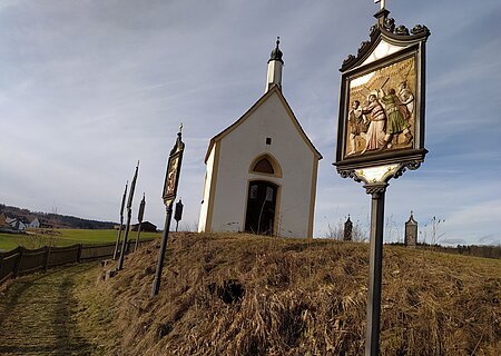 Kleine weiße Kapelle, die Kalvarienbergkapelle in Wittesheim, auf einem Hügel mit mehreren Kreuzwegstationen entlang eines Weges im Vordergrund.