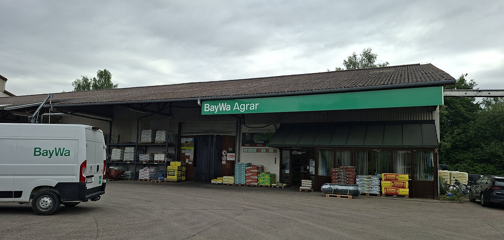 Außenansicht eines BayWa Agrar Gebäudes mit geparktem weißen Lieferwagen und Paletten mit Agrarprodukten davor.