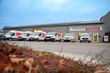 Firmengebäude vom Kachelofenbau mit sechs weißen Lieferwagen davor, am Gebäude dahinter ein Werbeschild „Martin Meyer Kachelofenbau“ und „Wir haben geöffnet“-Schild am Eingang. Vor den Lieferwagen eine Straße und unscharf ein bepflanztes Beet.