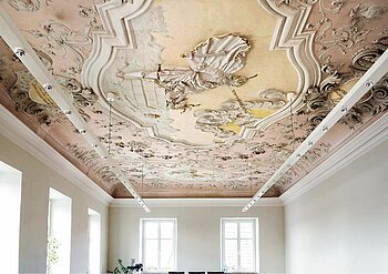 Farbig gestaltete Decke mit barocker Stuck- und Freskoverzierung in einem hellen Raum mit weißen Wände, vielen Fenstern und moderner an der Decke hängender Beleuchtung.