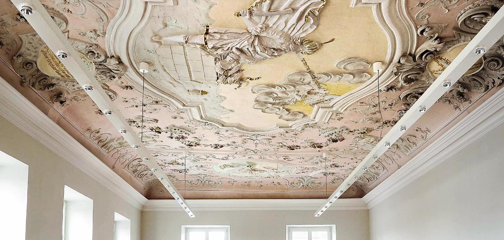 Farbig gestaltete Decke mit barocker Stuck- und Freskoverzierung in einem hellen Raum mit weißen Wände, vielen Fenstern und moderner an der Decke hängender Beleuchtung.