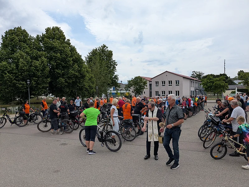 Große Gruppe von Radfahrern mit ihren Fahrrädern auf einem Parkplatz, ein Stadtpfarrer mit liturgischem Gewand segnet die Gruppe. Neben ihm ein weiterer Mann mit einem Behälter mit Weihwasser.