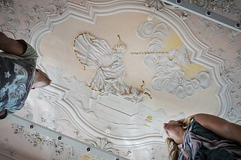 Stuckdecke im Monheimer Rathaus mit barocker farbiger Stuckreliefdarstellung und zwei Personen, die von unten darauf schauen.