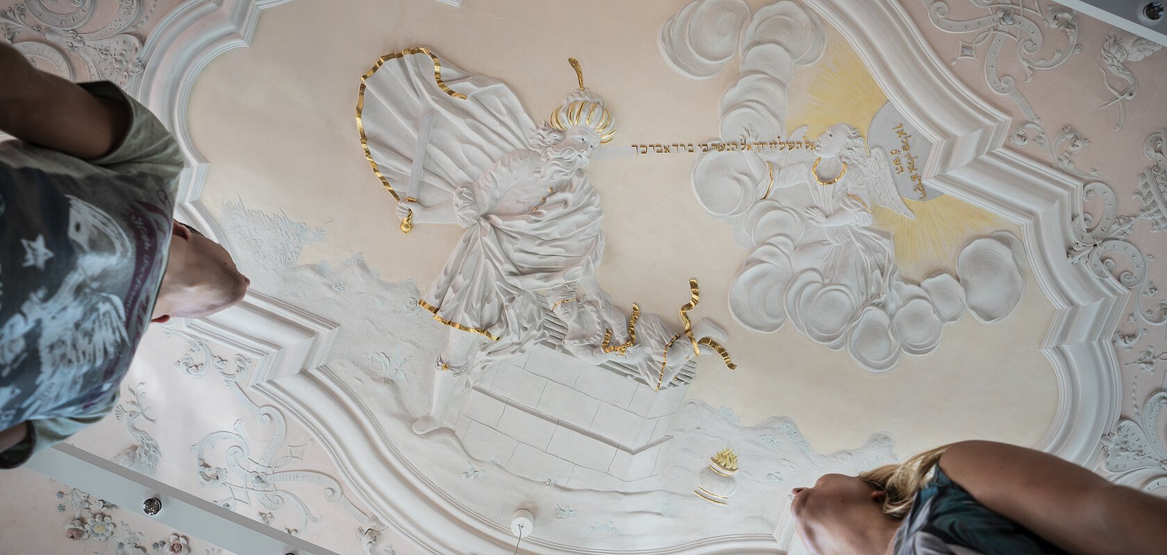 Stuckdecke im Monheimer Rathaus mit barocker farbiger Stuckreliefdarstellung und zwei Personen, die von unten darauf schauen.