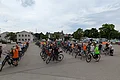 Viele Menschen mit Fahrrädern stehen auf einem großen Parkplatz vor Häusern und Bäumen unter bewölktem Himmel.