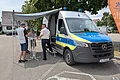 Polizeieinsatzfahrzeug mit ausgefahrener Markise und drei Personen an Stehtischen auf Parkplatz neben Bäumen im Freien. Im Hintergrund Wohnhäuser.