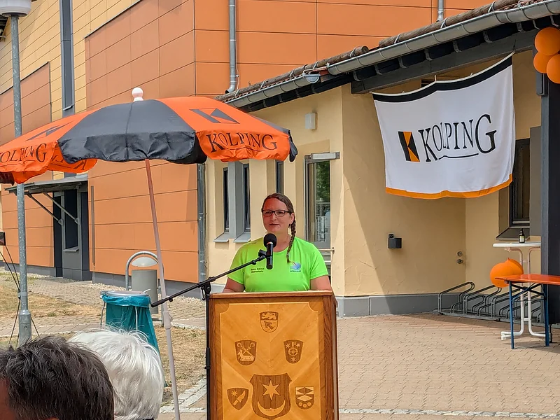 Frau im knalligem grünem T-Shirt spricht am Rednerpult aus Holz mit 9 verschiedenen Wappen unter einem Kolping-Schirm in orange-schwarz. Rechts ein Banner am Gebäude mit dem Schriftzug "Kolping" sichtbar, sowie orange Luftballons. Davor zwei Personen von Hinten mit Kolping-Monheim-Shirts.