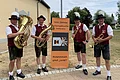 Vier Männer in Trachten mit Blasinstrumenten stehen links und rechts um einen Banner mit dem Text "100-Jahre Kolpingsfamilie Monheim - darunter das Logo vom Kolping - und weiter "Zusammen sind wir". Im Hintergrund eine Wiese mit Bäumen.