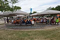 Menschen sitzen unter großen grauen Sonnenschirmen an Tischen auf Bänken im Kreis um eine Statue im Freien. Im Vordergrund eine Wiese bei bewölktem Himmel.