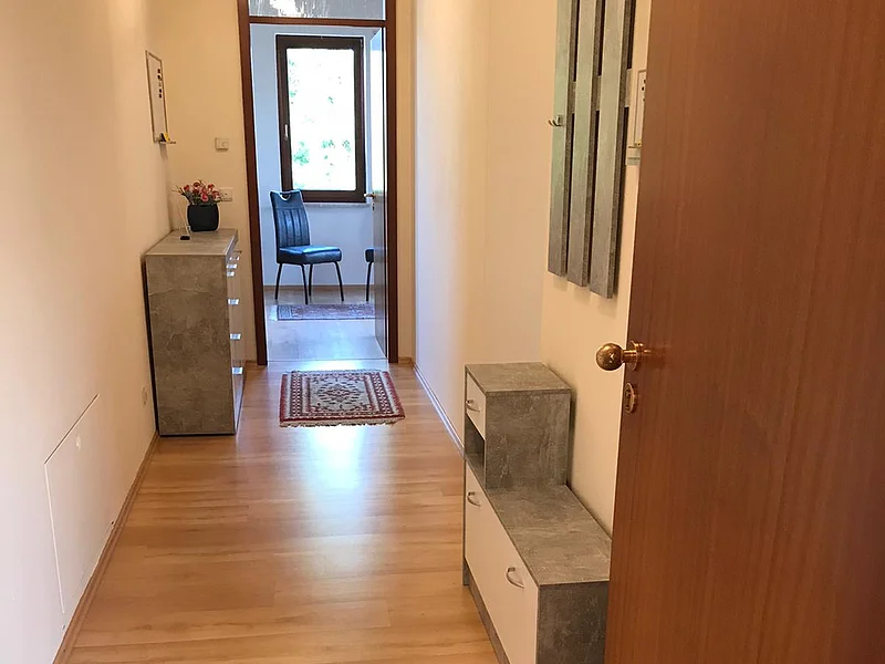 Wohnung zur Vermietung - Monheim, Am Sonnenbühl 5 Flur mit Holzfußboden, grauen Möbeln, kleinem Teppichen am Ende des Flurs und Blick in einen Raum mit Fenster und Stuhl.
